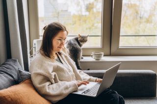 A woman sat on a sofa working on a laptop next to a cat