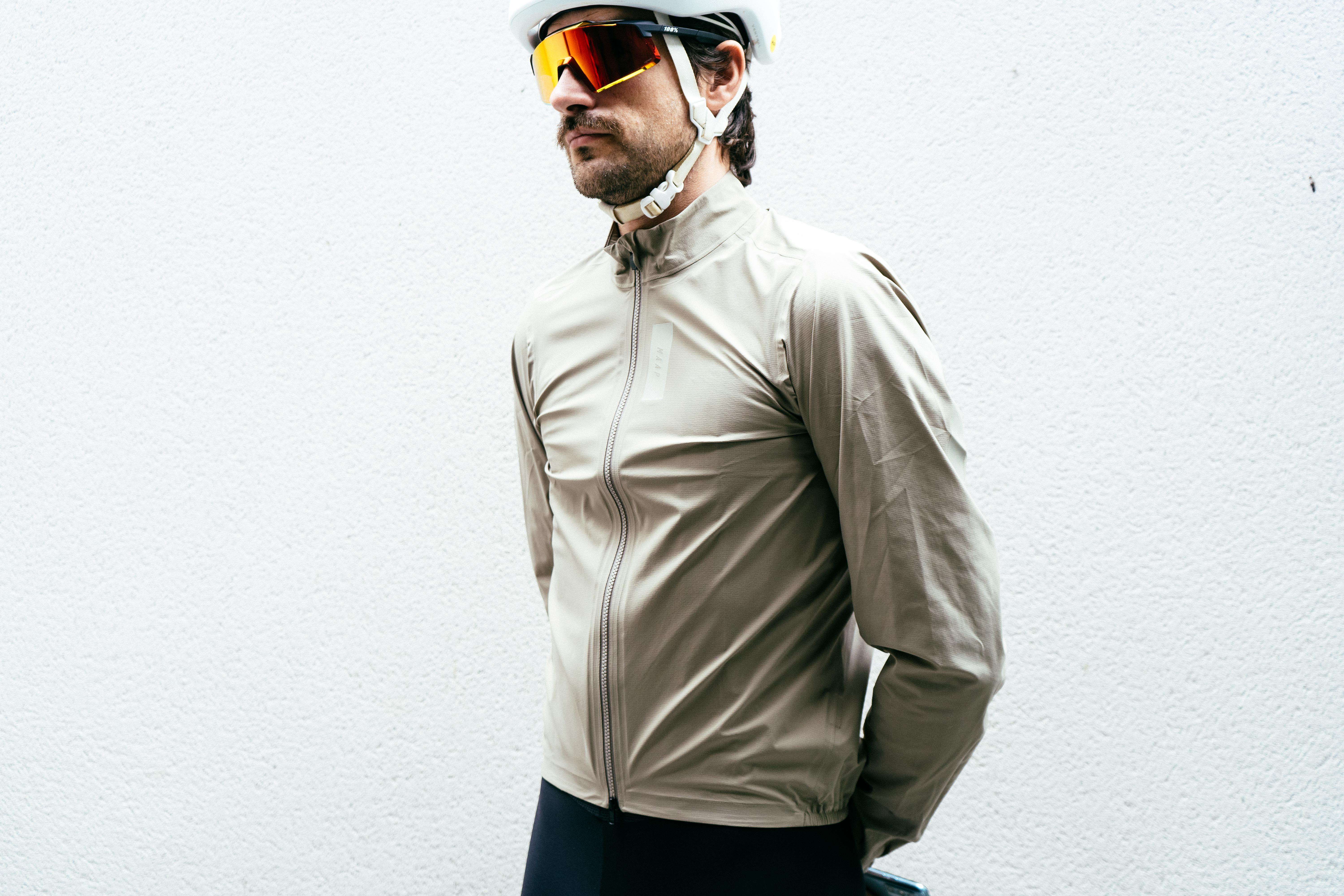 A man in an off-white cycling rain jacket, white helmet, and orange sunglasses against a white wall