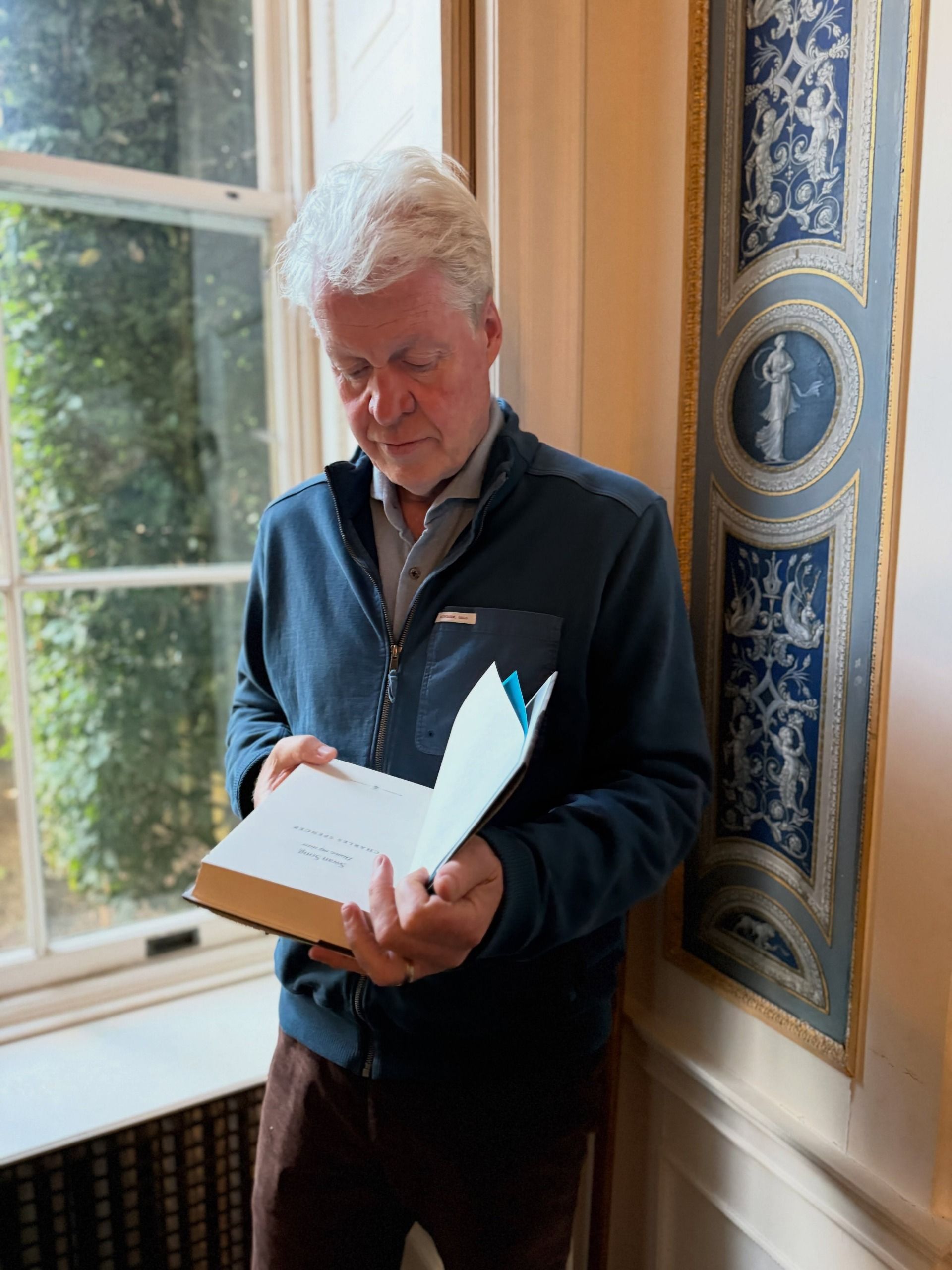 Earl Spencer reading a copy of his book Swan Song by a window