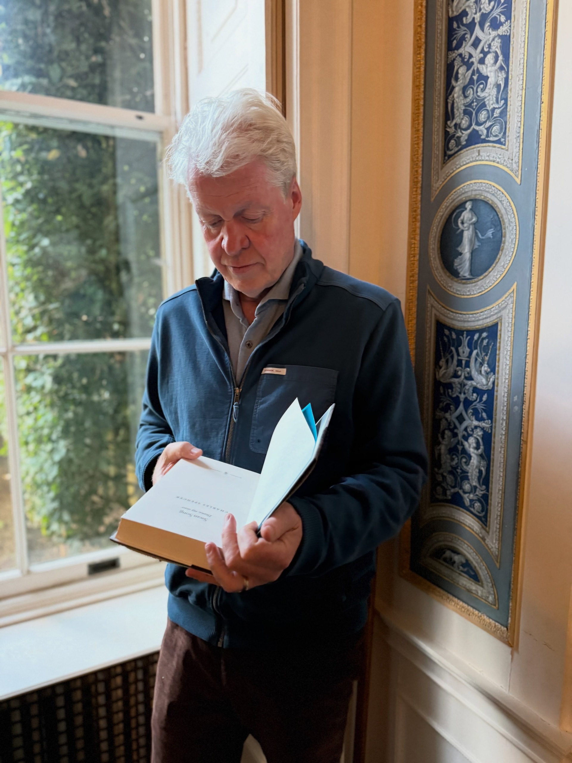 Earl Spencer reading a copy of his book Swan Song by a window
