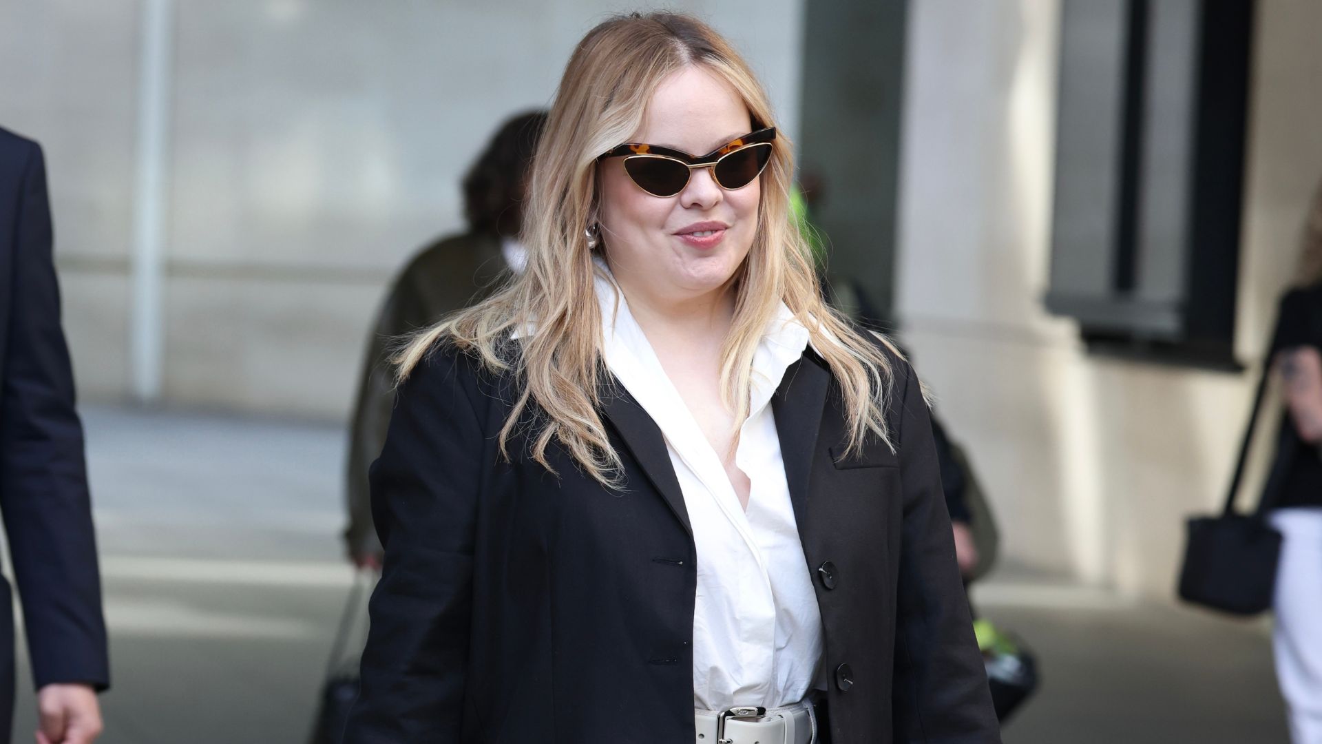 Nicola Coughlan walks down the street wearing white jeans with silver platform heels, a white shirt and a black blazer.