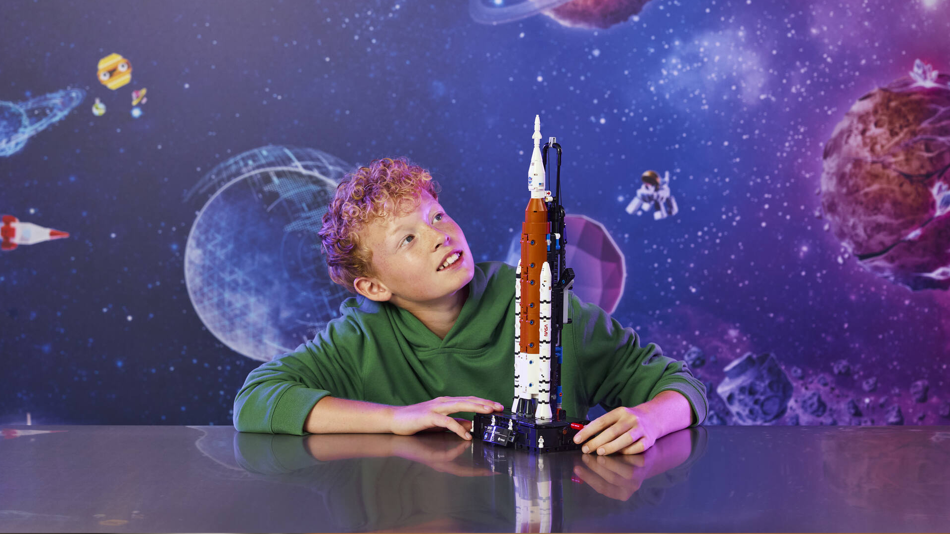 A child smiles while looking at a NASA rocket set, with a cosmic background visible behind them