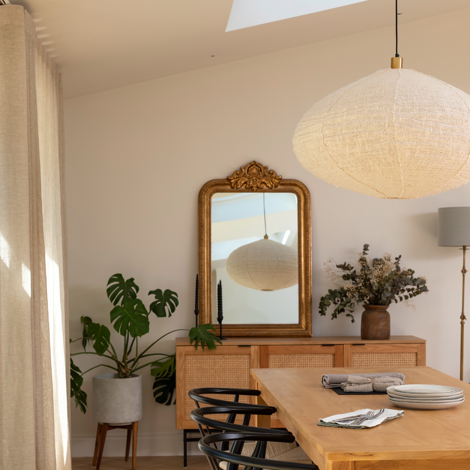 A white-painted dining room with a wooden dining table, a rattan sideboard with a mirror and a paper lantern pendant lampshade