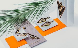 Geometrically crafted jewelry pieces that shimmer with hints of mirrored gold, bright orange and blue on orange and wooden panels with a palm leaf next to them.