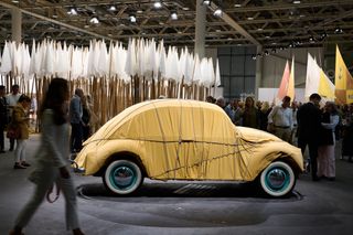 A car wrapped in a yellow fabric sits at the center of a filled-with-installation gallery space.