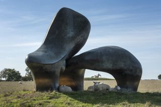 A two-part, gigantic bronze sculpture with an abstract shape is surrounded by a herd of sheep in a sun-filled field.