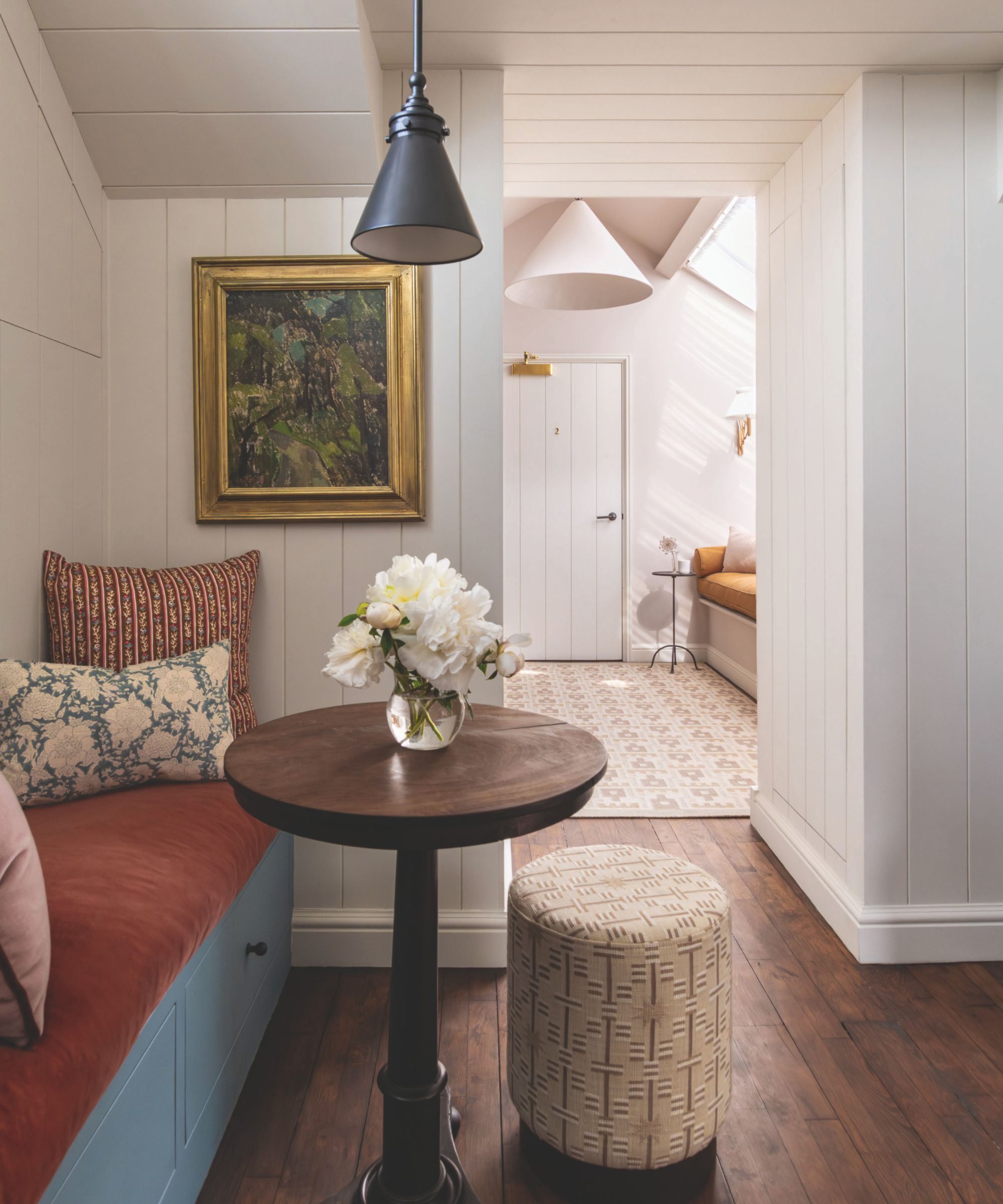 An interior photograph of a small dining nook with light grey wood paneling, a rust velvet banquette with patterned pillows, a round wooden table, and a patterned ottoman.