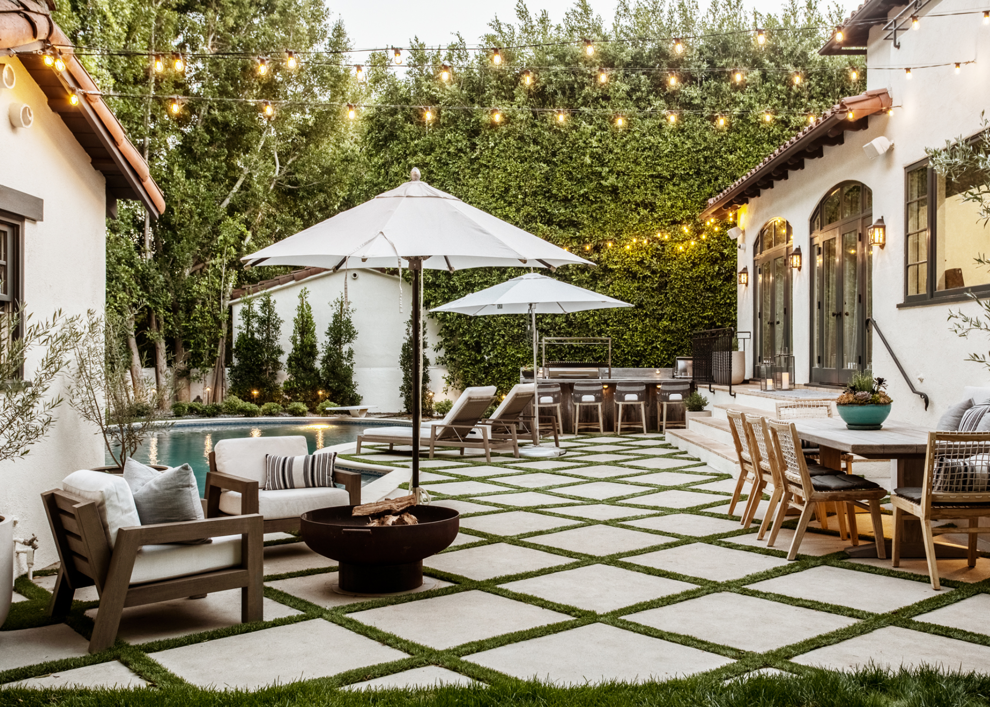 An outdoor space with string lights and patio furniture as well as parasols. There is a large dining table on one side of the garden