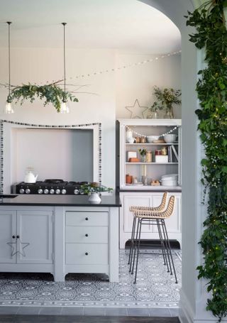 A grey and white Christmas-themed kitchen with two ceiling lights with seasonal foliage, white dresser, wire star decor and patterned floor