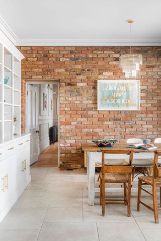 exposed brick wall in kitchen diner with old farmhouse table and chapel style wooden chairs and white bookcase