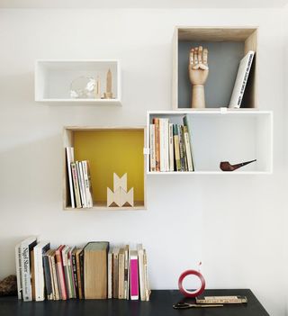 Mini Stacked Shelf, four white cubes and rectangle box shelves on a wall containing a selection of books and items, a black surface underneath also containing a selection of books