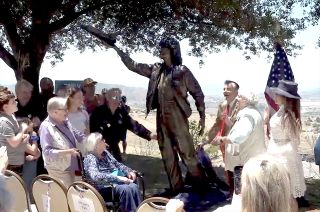 a crowd of people admire a bronze statue of sally ride with a tree and an american flag in the background