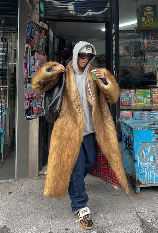 A woman walking out of a convince store wearing a gray hoodie over a baseball hat with a brown fur coat, cargo denim, and cheetah print sneakers.