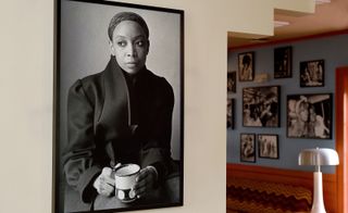A large black & white photo of a black female is hung on the wall to the left. Through an opening to the right, we see black & white photographs of black female models hung on a light blue and white dotted wall.