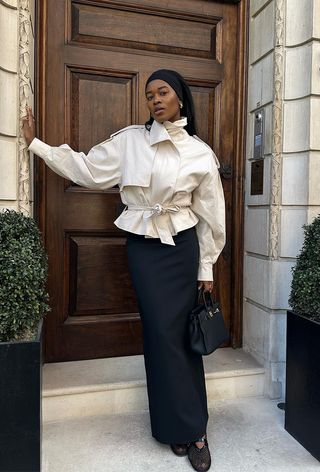 a black maxi skirt outfit shown on a woman wearing a black headband, silver earrings, a tan cropped trench styled with a black maxi pencil skirt and black mesh flats