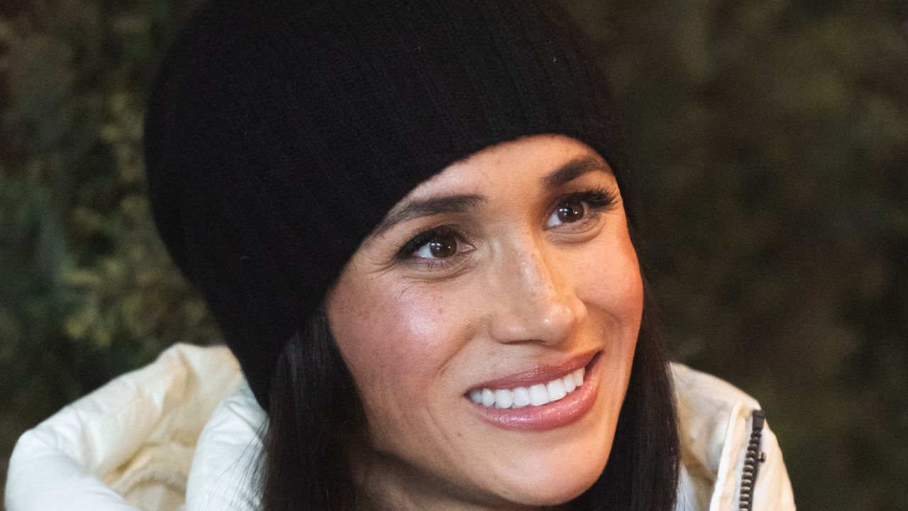 Meghan Markle wearing a black winter hat and a white puffer coat smiling and looking to the right