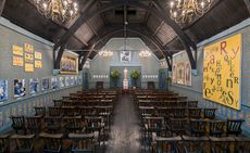 The Oscar Wilde Temple, rows of wooden chairs on either side of an isle leading to an Oscar Wilde cream statue with planted trees either side, dark wooden floor, arched wooden ceiling, four lit chandeliers, decorated walls with colourful artwork 