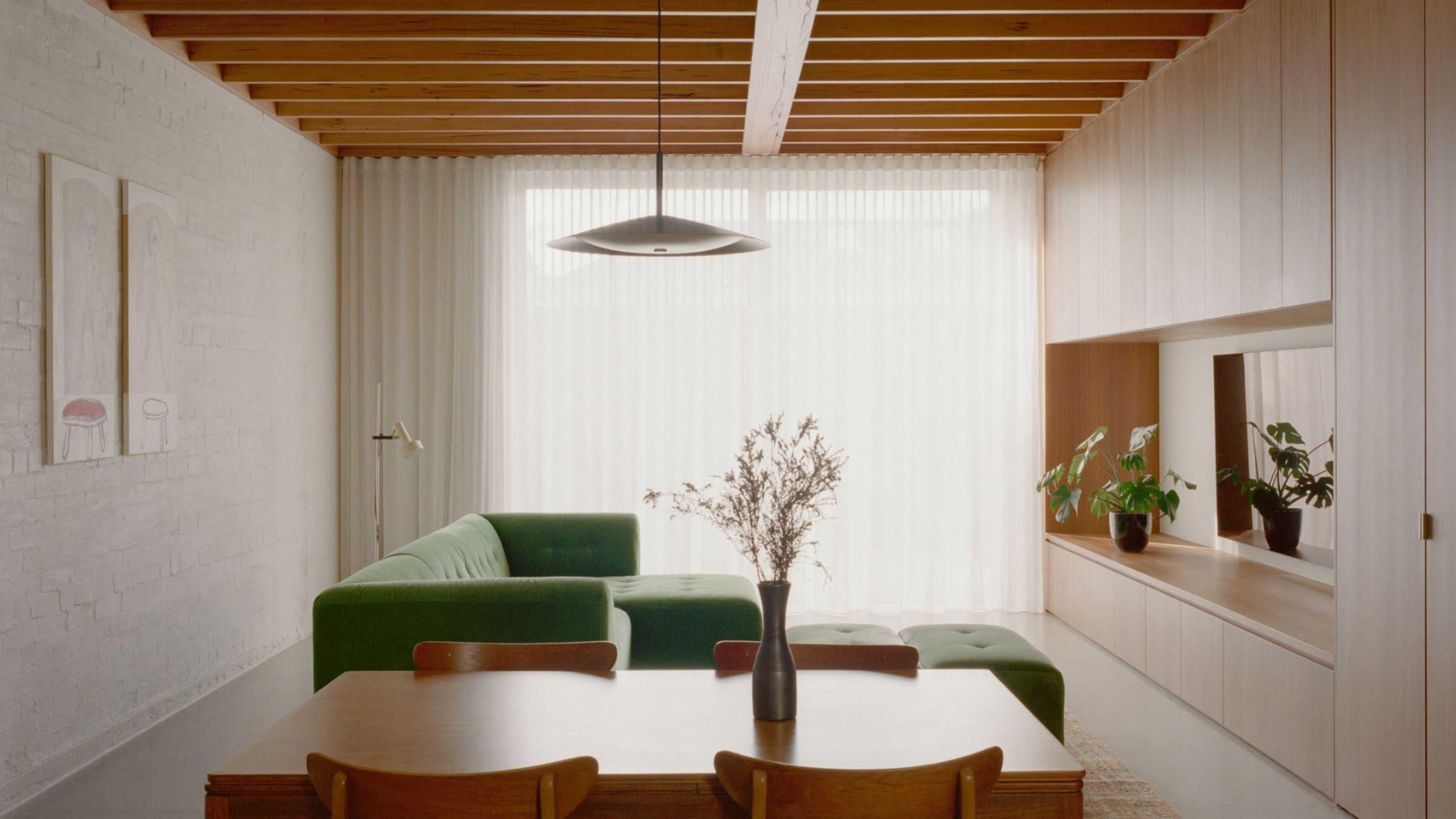 modern living room with white rendered brick wall, timber joinery around TV with plant pot, sheer curtains pulled across windows, green sofa floating in the room, and timber dining table