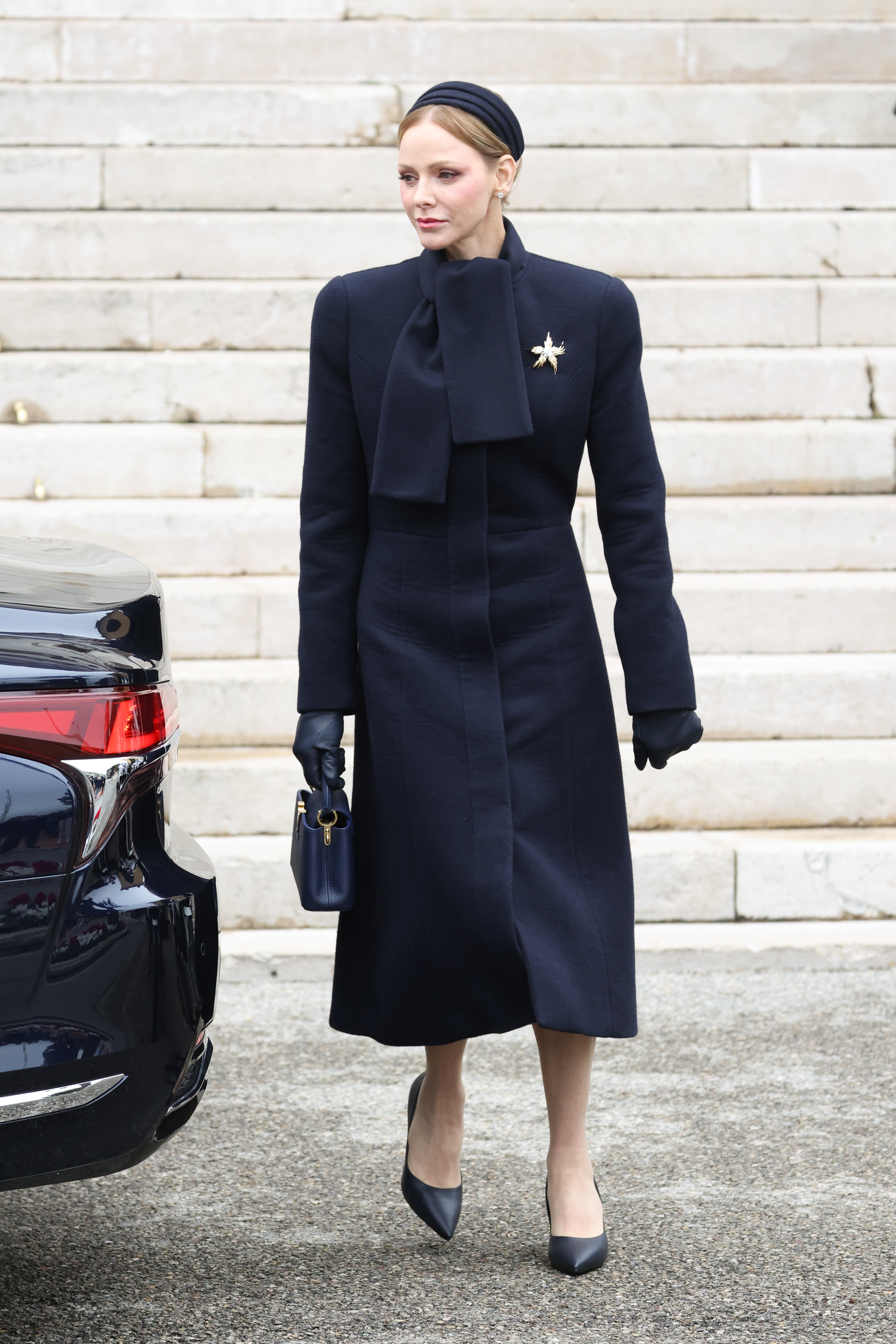 Princess Charlene wearing a long navy coat walking in front of a car
