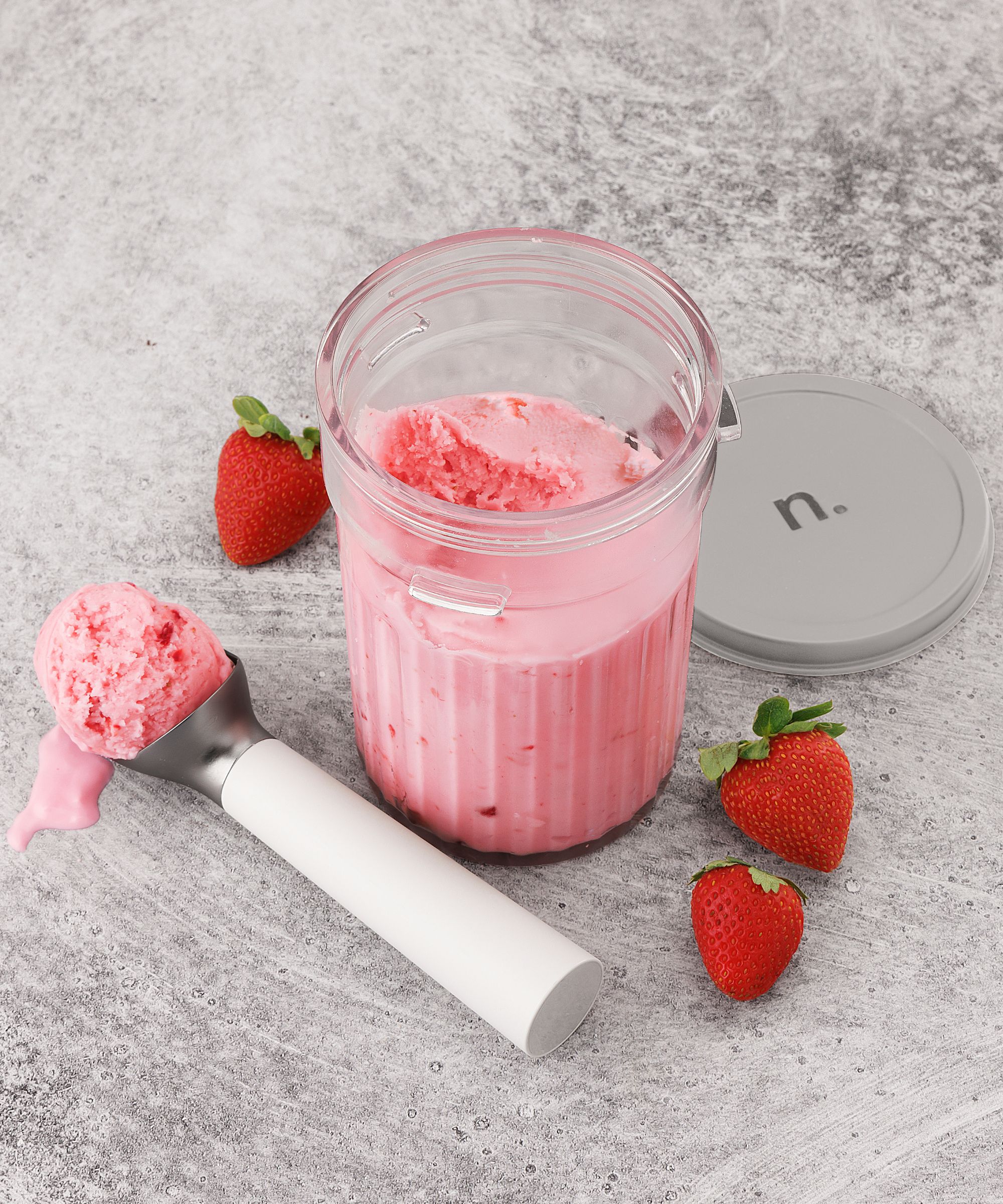 A clear container filled with pink strawberry ice cream, with a scoop beside it with a ball of ice cream in it. Three strawberries on the counter beside it.