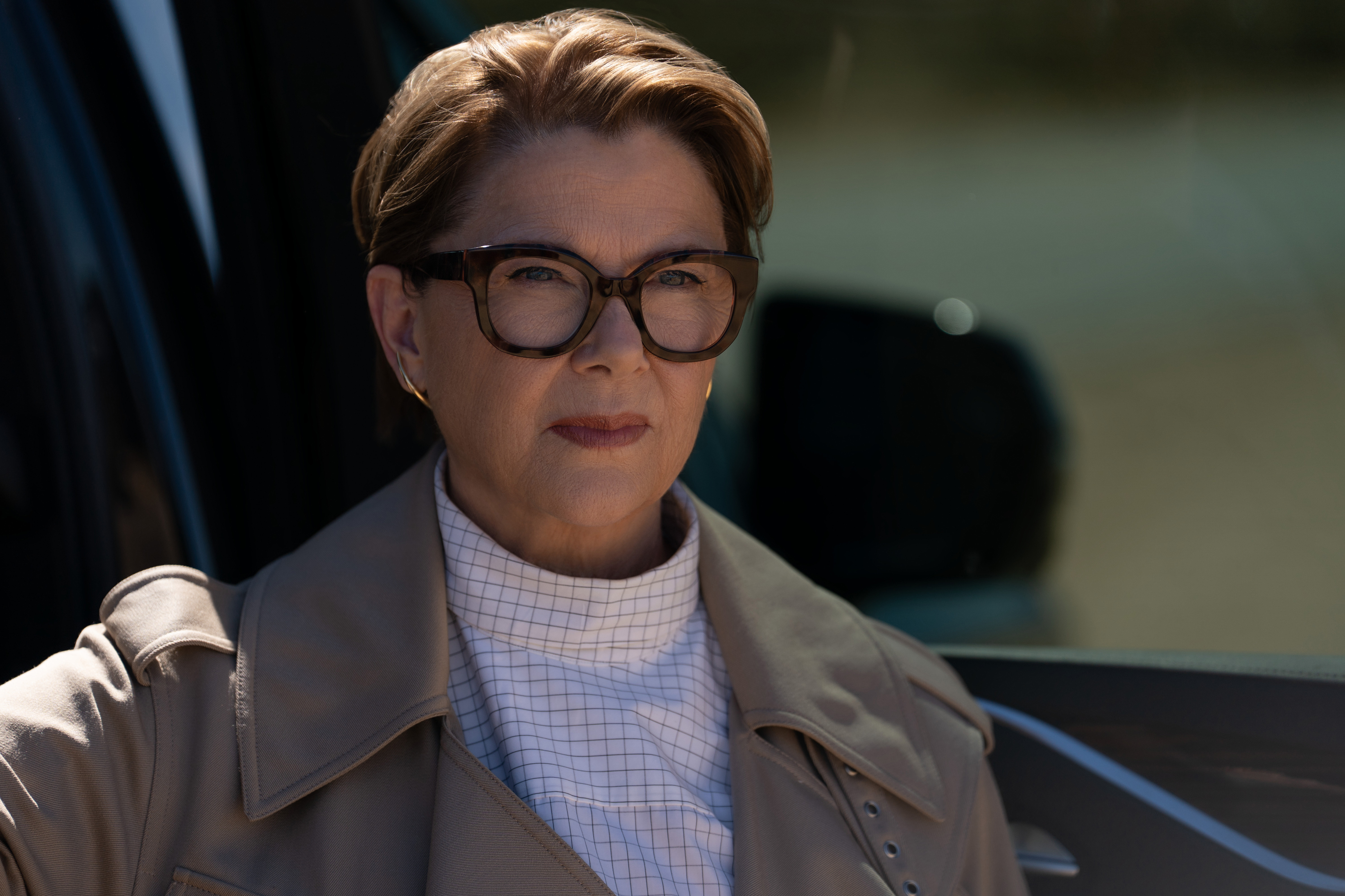 annette bening wearing glasses and a trench coat standing outside of a car in a still from apple tv's lucky