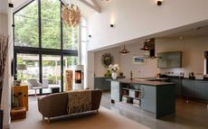 An open plan kitchen and lounge area with a kitchen island and counter with grey cupboards, a brown sofa and a grey chair next to large windows.