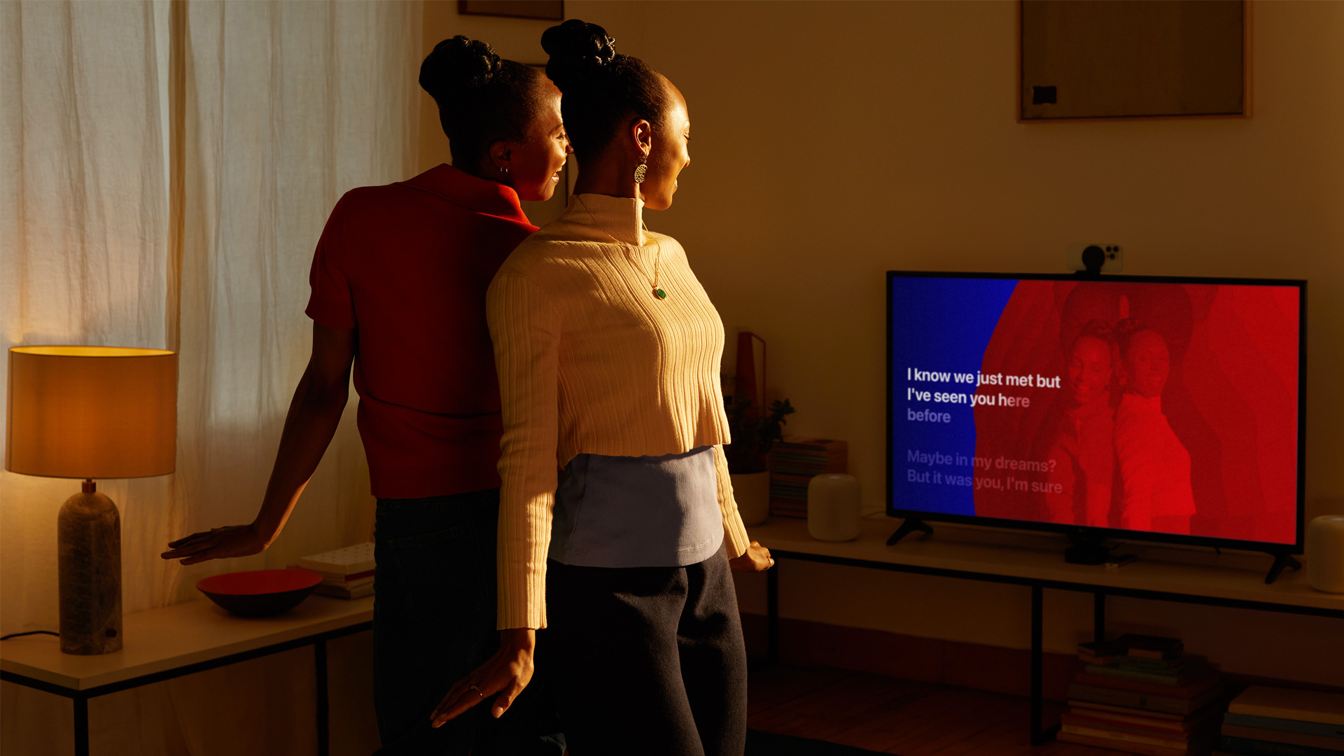 Two friends stand in front of a TV singing along to lyrics with Apple Music Sing