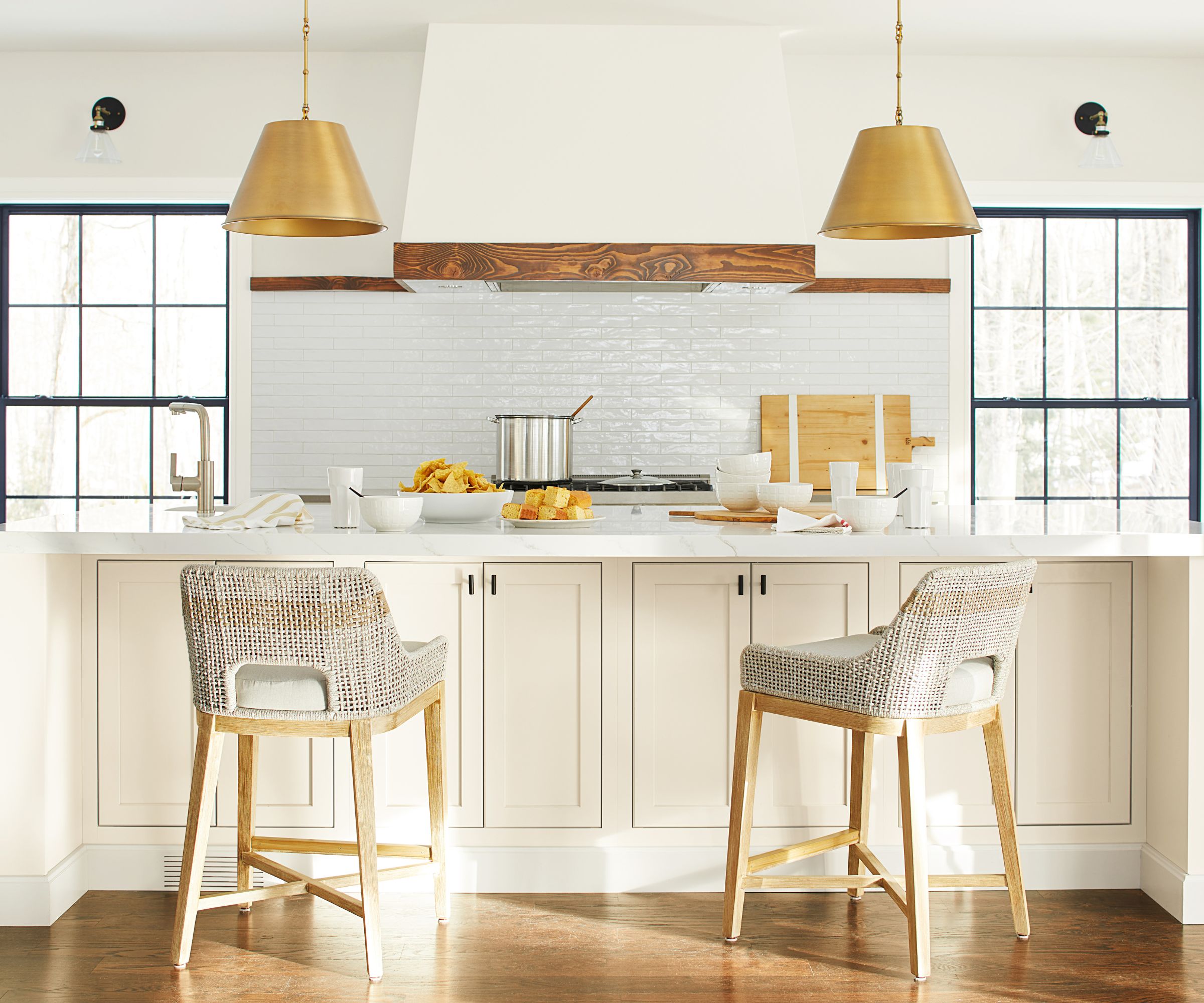 White kitchen with white subway tiled backsplash, two large windows with black frames, a cream island with a white countertop, two gold pendant lights hanging above, and two wooden bar stools.