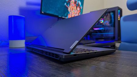 A Lenovo Legion 5i (Gen 9) gaming laptop on a desk with a blue backlight, facing away at an angle with its lid open to show its rear and right side ports, a bit of its RGB-lit keyboard, and the "Lenovo" logo on the lid.