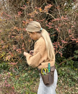 Woman wearing brown leather gardening tool belt holding two pairs of pruning shears, whilst pruning viburnum in fall