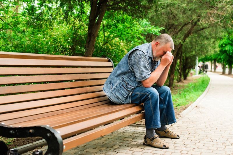 An older man sits on a park bench, looking very sad.