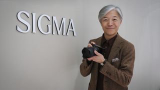 Sigma CEO Kazuto Yamaki, holding a Sigma BF, in front of the Sigma logo at CP+ 2026