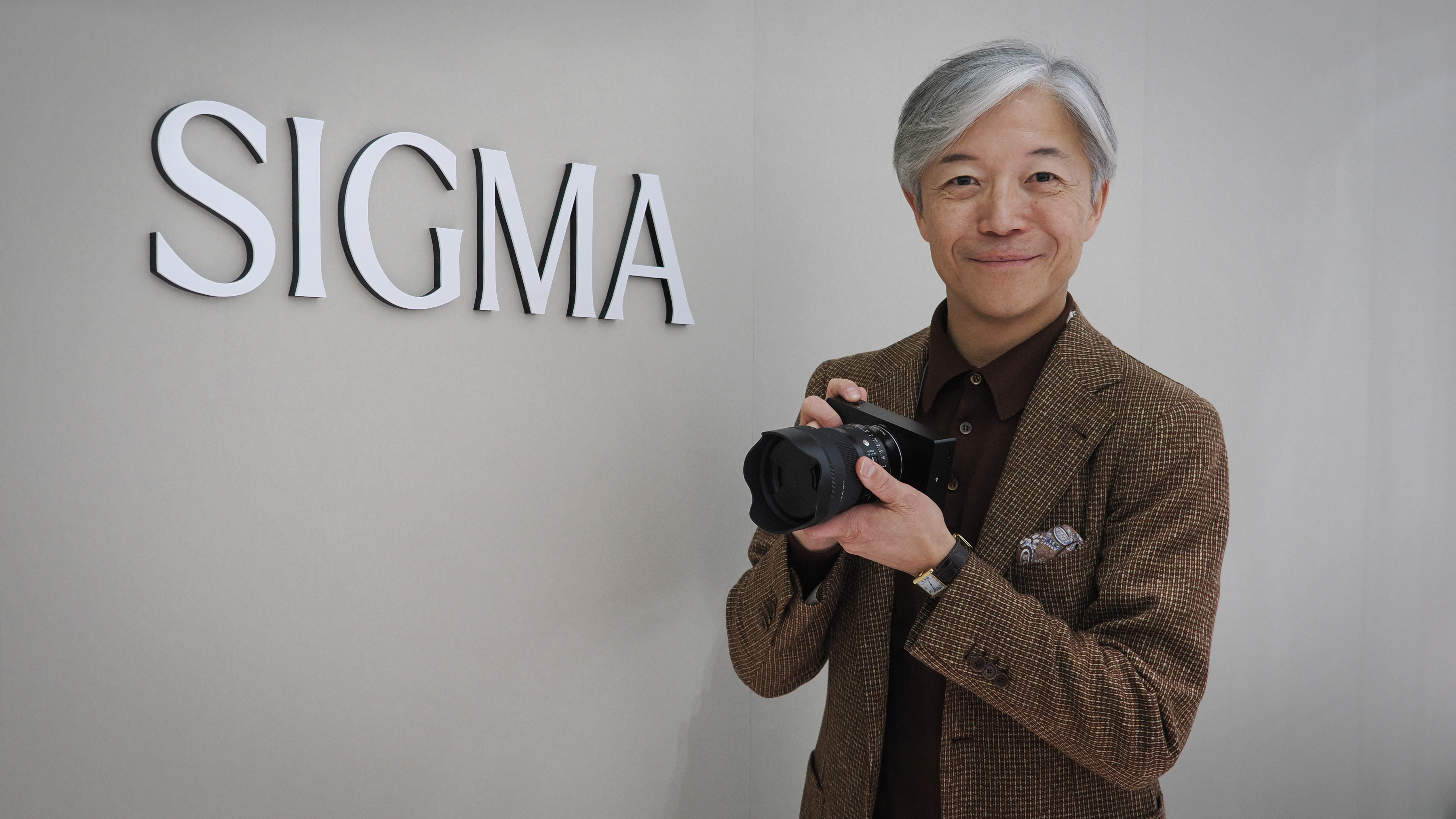 Sigma CEO Kazuto Yamaki, holding a Sigma BF, in front of the Sigma logo at CP+ 2026