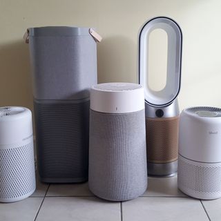 Five air purifiers of various sizes lined up on a tiled floor in a room with a pale yellow wall