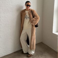 A woman wears a faux-fur coat, crew neck graphic sweater, white sweatpants and suede trainers.