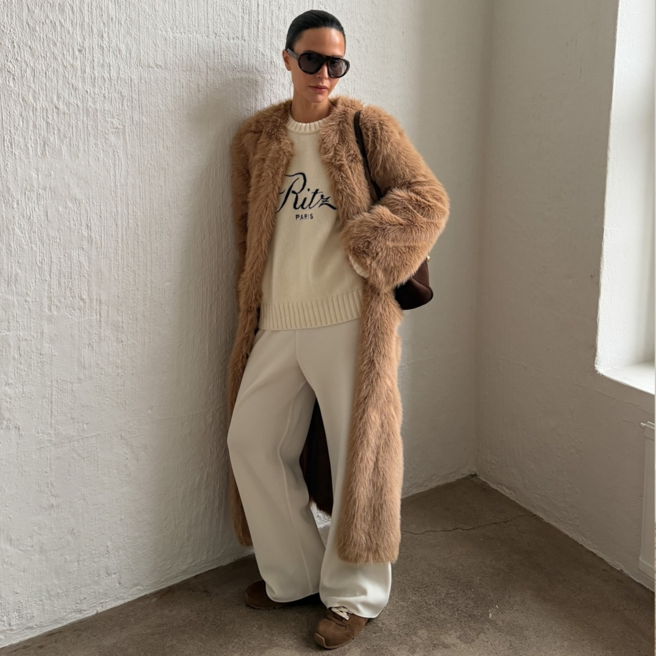 A woman wears a faux-fur coat, crew neck graphic sweater, white sweatpants and suede trainers.