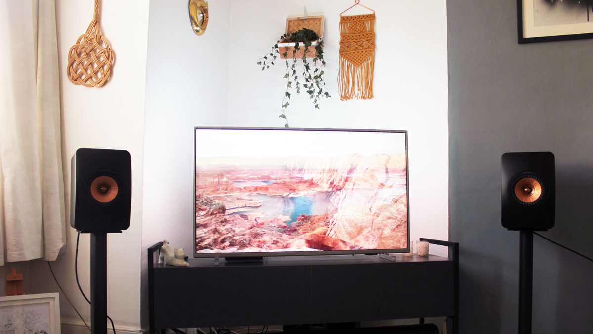 KEF LS50 speakers sit on each side of a television, which shows an American canyon landscape. The bronze speaker driver of the Kef speakers is visible in contrast to their dark body overall