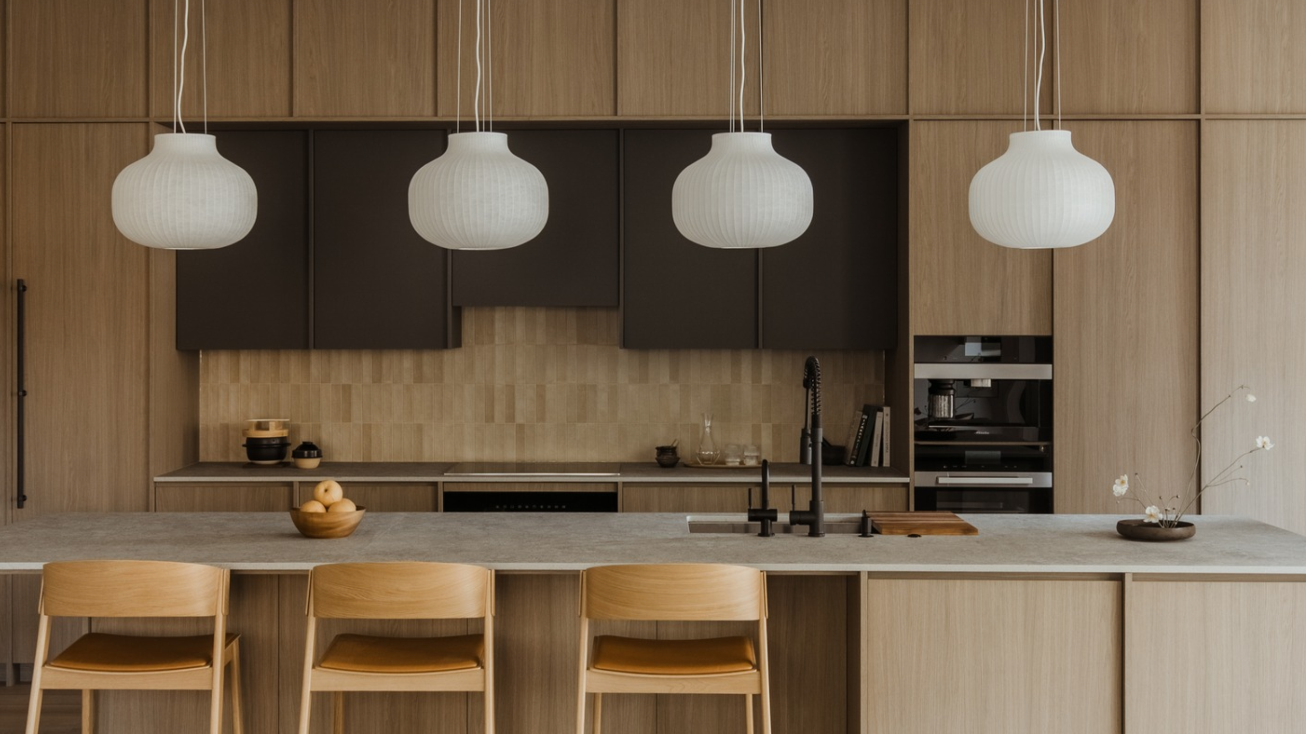 A minimalist Japanese kitchen with a row of lantern-like pendants above the island, wooden cabinets, three stools, a bowl of fruit, and an ikebana arrangement