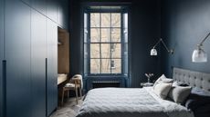 dark navy blue bedroom with large window, radiator under it, built-in blue cabinetry with a timber desk built-in and timber chair, color drenched walls, two wall sconces on either side of the bed, gray headboard, blue and gray sheets