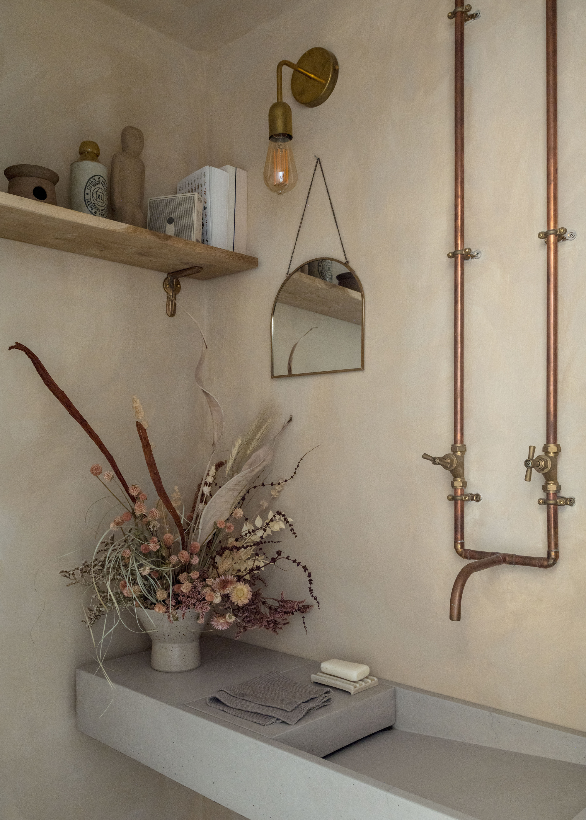 A beige limewash bathroom with brass pipes and taps, a gray sink with a vase of dried flowers, a hanging mirror, a wall sconce and a shelf of bath products