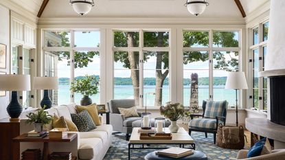 Large sunroom with three sets of double doors looking out onto the sea, cream walls, a large fireplace, a cream sofa, a blue floral rug, a decorated coffee table and lots of upholstered seating.