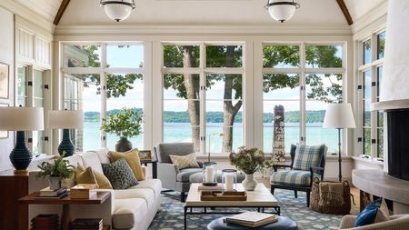 Large sunroom with three sets of double doors looking out onto the sea, cream walls, a large fireplace, a cream sofa, a blue floral rug, a decorated coffee table and lots of upholstered seating.