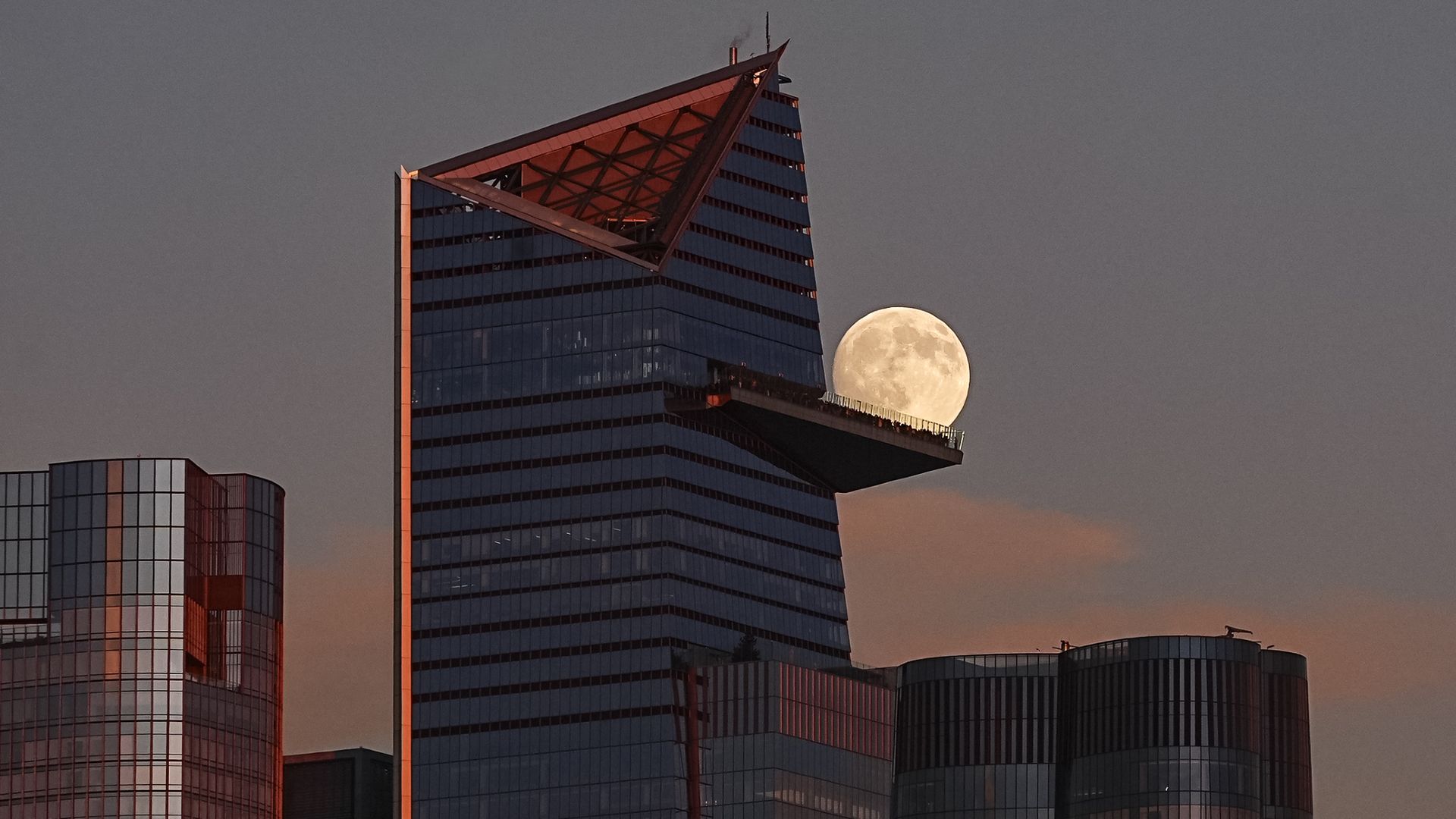The moon is photographed shining close to the observation deck of a skyscraper in a darkening sky.