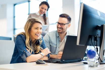 A young woman and man laugh in front of their office computer screens.