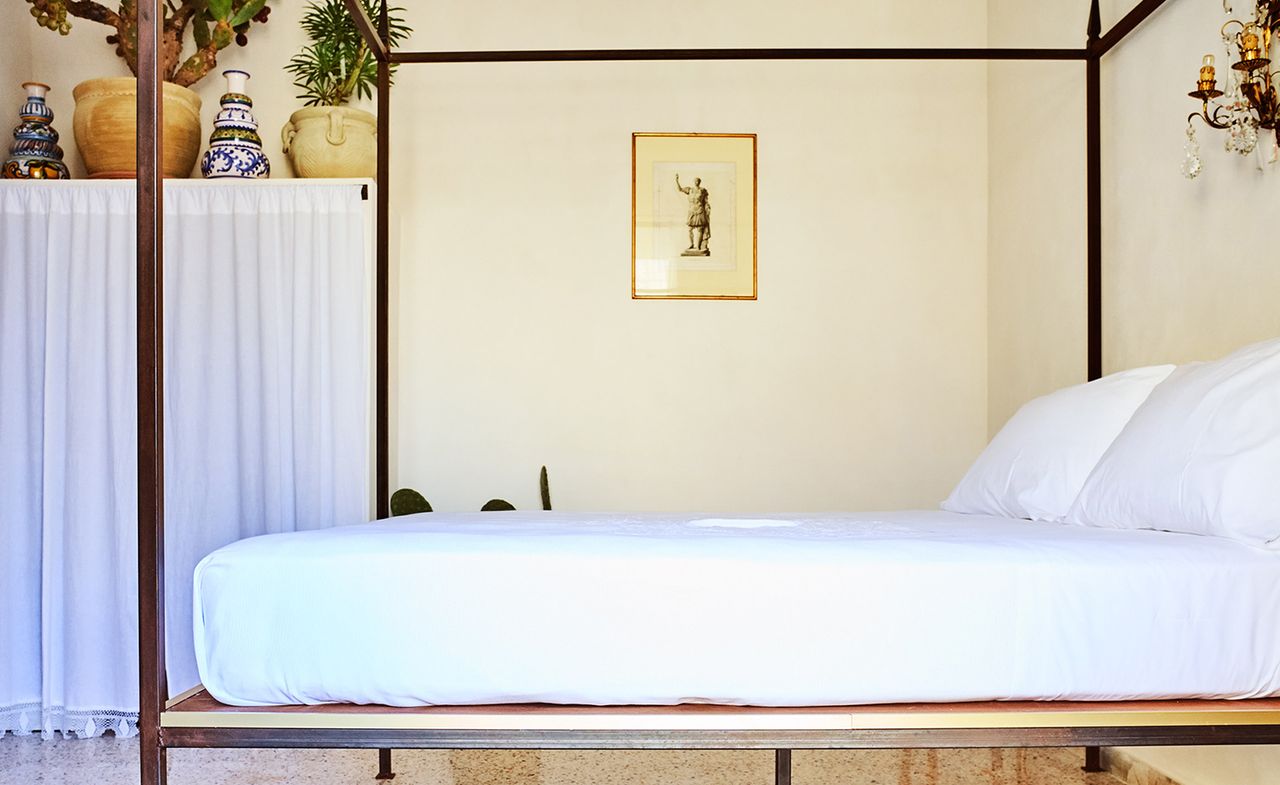 A hotel room with a four post bed, a cupboard with decoration on and a wall painting.