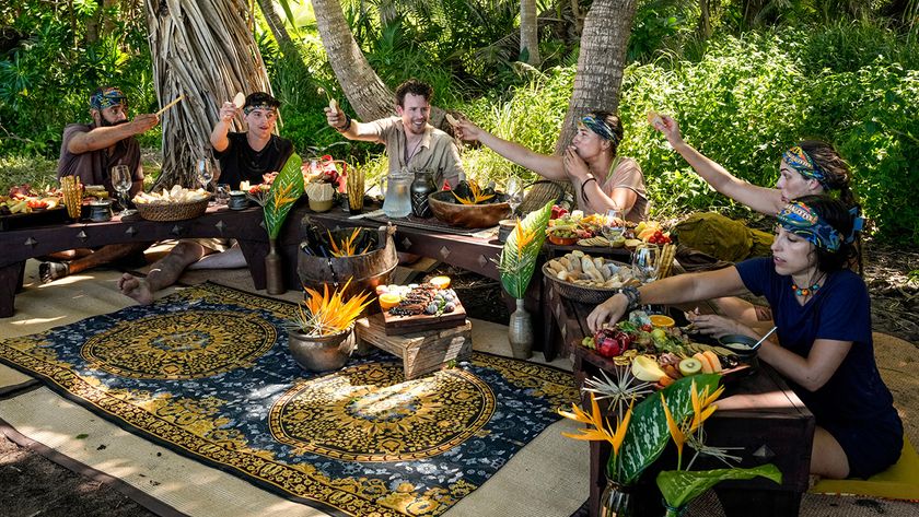 The cast of Survivor Season 49 sit on rugs around a low seated table with an abundance of food, raising a toast.