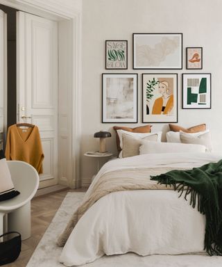 A white bedroom with a gallery wall with six prints, a white bed with four throw pillows, a white duvet and beige and dark green throws, a rug underneath the bed, and a chair and door with an orange shirt hung up