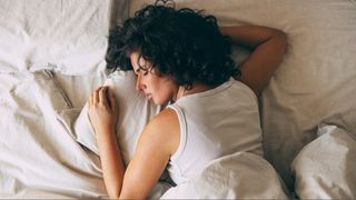 Woman with black curly hair wearing a white vest top sleeping on her front in bed with white sheets.