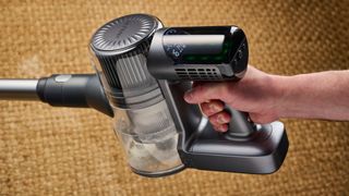 the handle of a modern-looking sleek shiny vacuum cleaner with light grey components on a carpet in front of a blue background