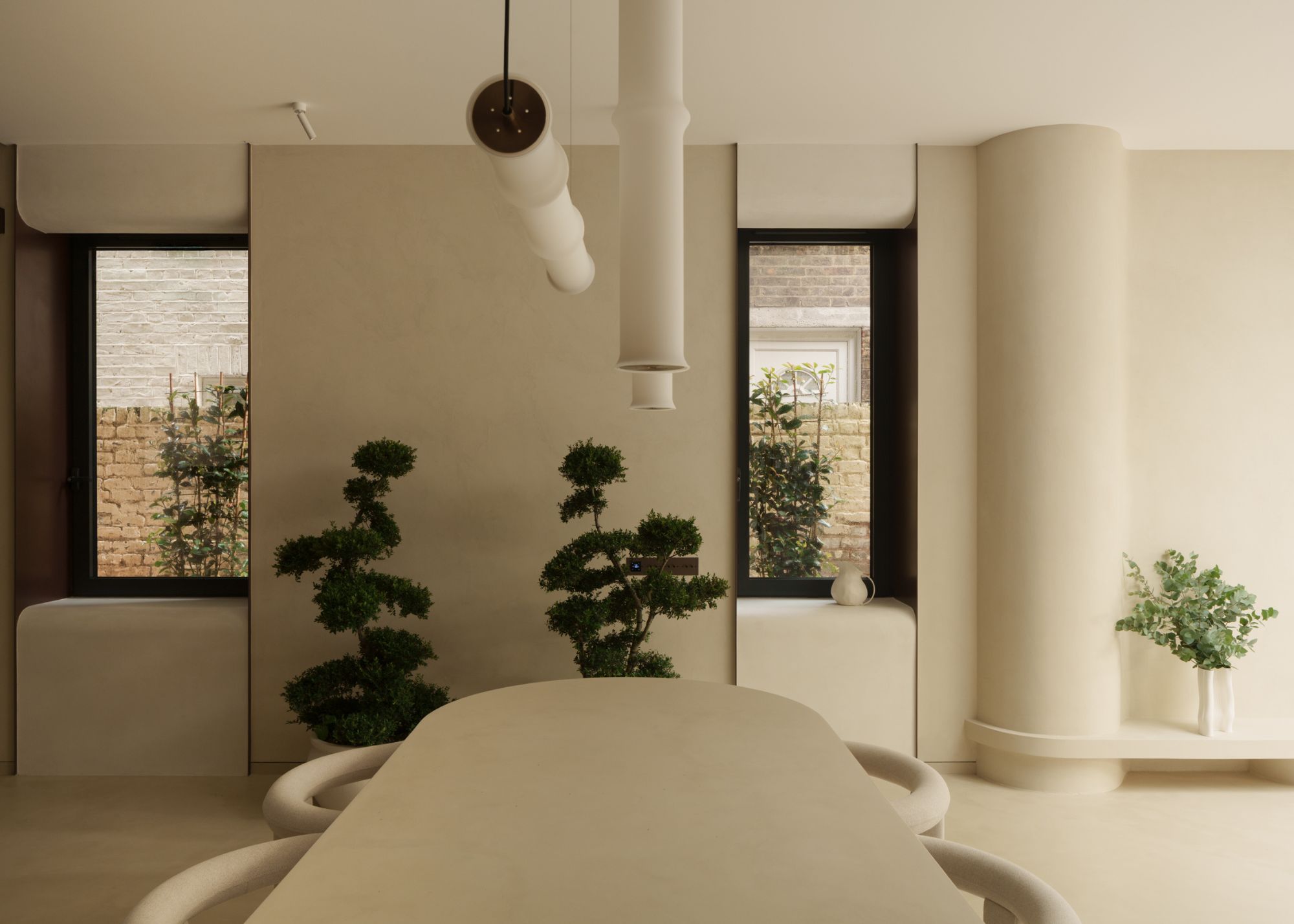 A neutral dining space with a cream oval dining table and Japanese cloud plants against the wall between two windows, with microcement finishes all around